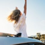 Retrato de uma mulher com metade do corpo para fora do teto solar do carro, com os braços erguidos, e tendo a estrada adiante