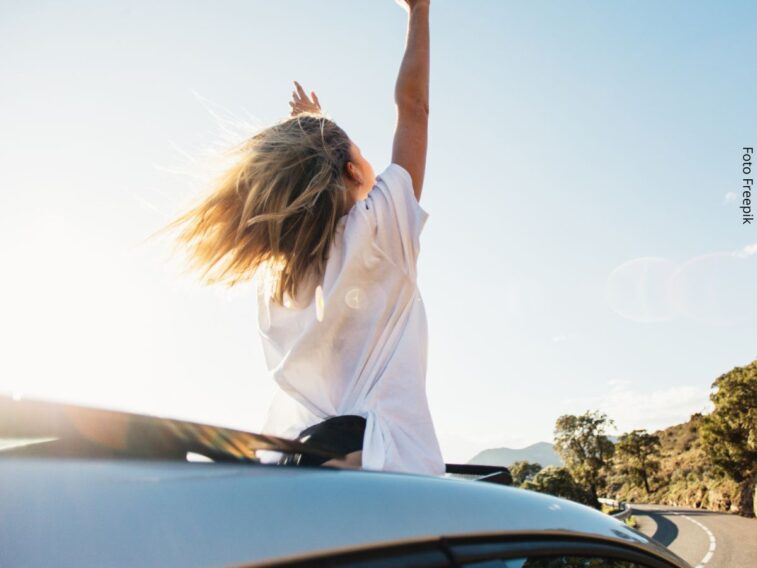 Retrato de uma mulher com metade do corpo para fora do teto solar do carro, com os braços erguidos, e tendo a estrada adiante