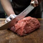 Homem corta carne vermelha com luva em uma das mãos, e usando faca e tábua exclusivas para evitar contaminação cruzada.