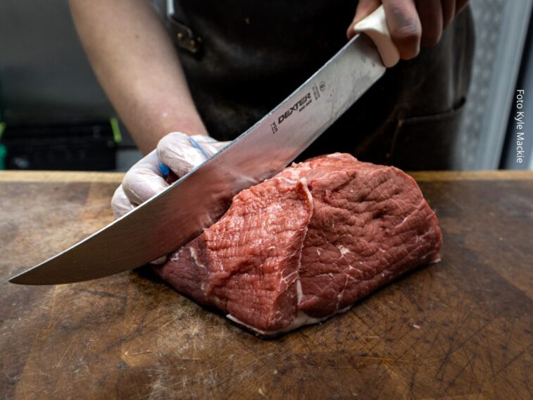 Imagem de um pedaço grande de carne vermelha sobre uma tábua, sendo cortada por um homem usando luva em uma das mãos para evitar a contaminação cruzada