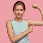 Retrato de uma mulher jovem e de aparência agradável com o dedo indicador apontado para o bíceps do braço esquerdo. Ideia de perda de massa muscular. Está usando camiseta azul claro, tendo ao fundo uma parede rosa