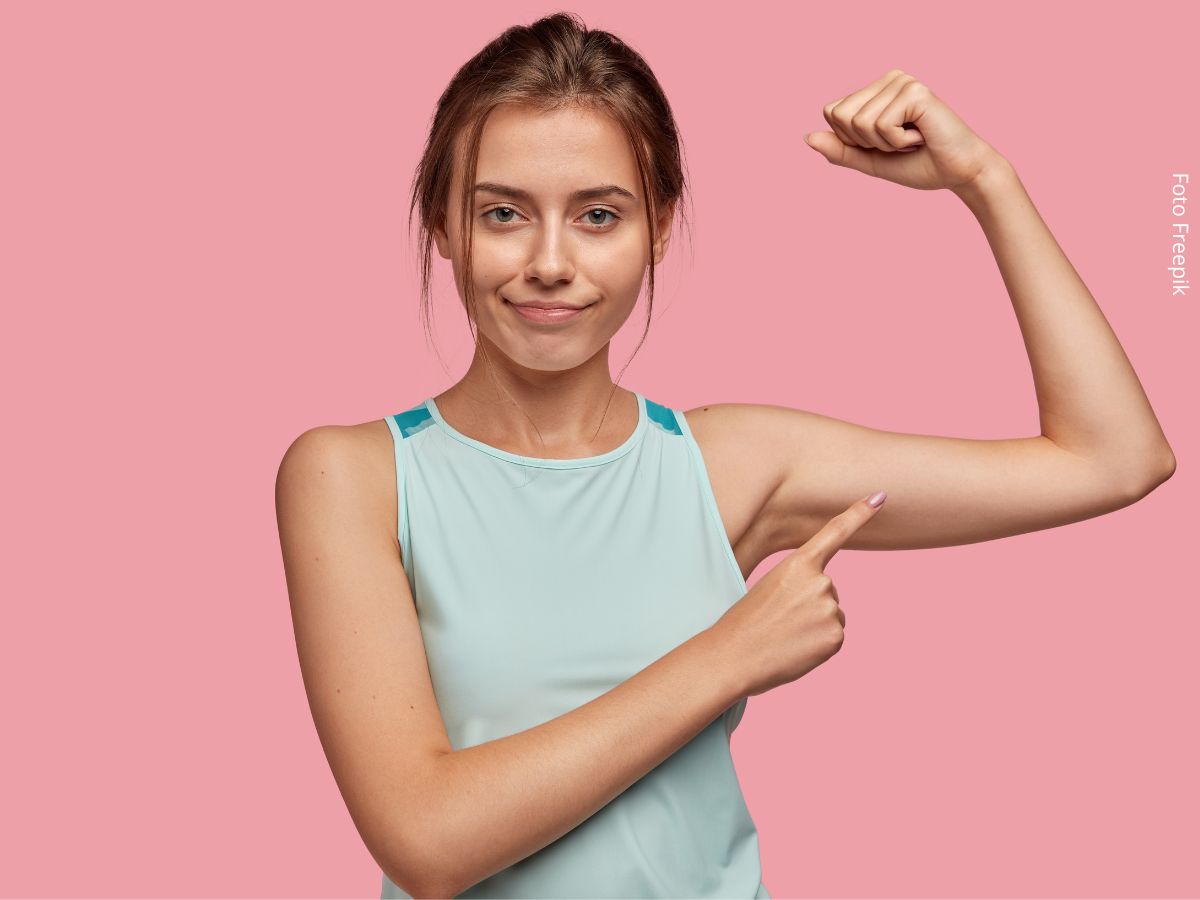 Retrato de uma mulher jovem e de aparência agradável com o dedo indicador apontado para o bíceps do braço esquerdo. Ideia de perda de massa muscular. Está usando camiseta azul claro, tendo ao fundo uma parede rosa