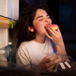 Mulher pega um doce na geladeira e começa a comê-lo antes de fechar a porta, representando o impulso causado pela fome emocional.