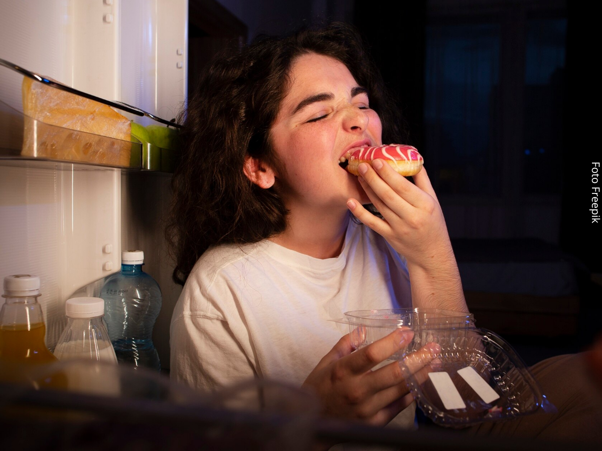 Mulher pega um doce na geladeira e começa a comê-lo antes de fechar a porta, representando o impulso causado pela fome emocional.
