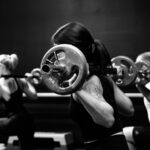 Imagem mostra mulher em primeiro plano fazendo agachamento com uma barra de de pesos sobre as costas, acompanhada de mais duas pessoas também se exercitando ao fundo
