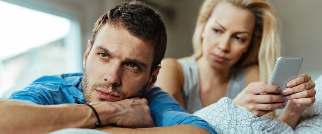 Imagem de um casal deitado na cama, em que o homem aparece pensativo, aparentemente distante da mulher ao lado. Ideia de relacionamento em crise