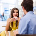 Imagem mostra mulher jovem sorrindo, segurando uma xícara de café sentada à mesa de frente para um homem que aparece de costas. Ideia de um início de relacionamento