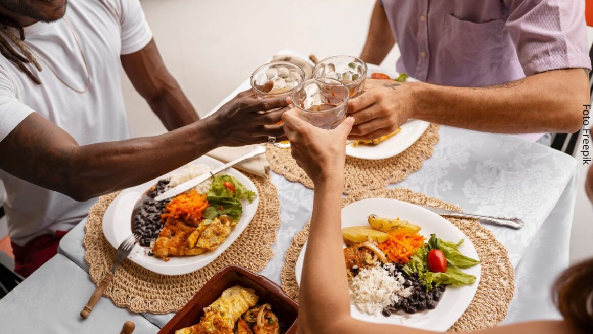 Foto de três amigos sentados à mesa para almoçar, e brindando com copos de cerveja na mãos. Ideia de alimentação intuitiva