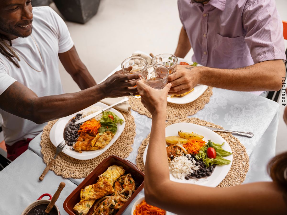 Foto de três amigos sentados à mesa para almoçar, e brindando com copos de cerveja na mãos. Ideia de alimentação intuitiva