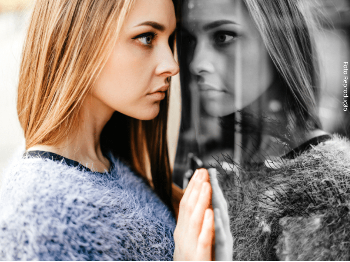 Close-up de jovem mulher de perfil, com expressão fechada, parada diante de uma vitrine de vidro refletindo seu rosto