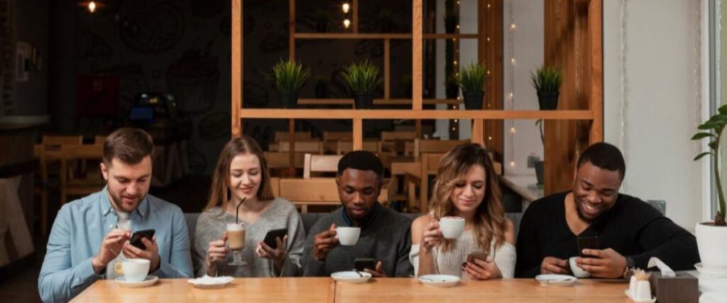 Cinco amigos sentados em uma mesa de restaurante, todos olhando para um celular, ilustrando a desconexão mesmo na presença física.