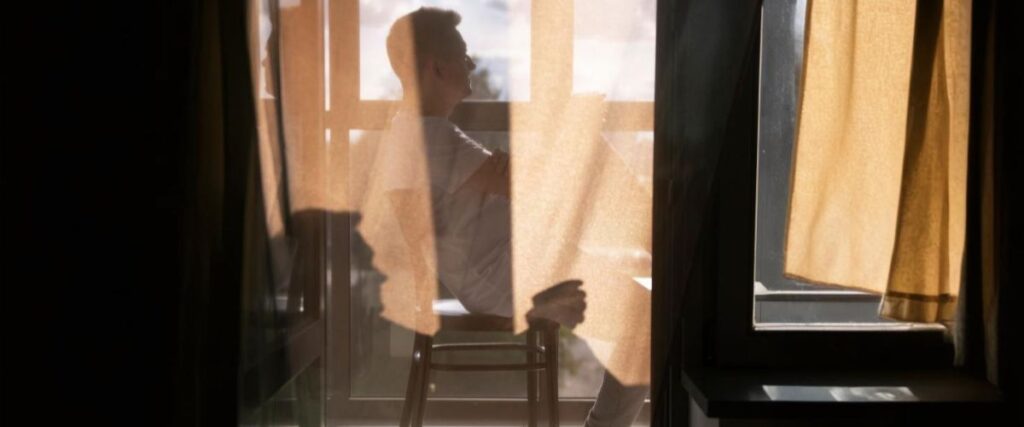 Imagem de um homem sentado na varando com uma xícara de café na nas mãos sereno olhando a paisagem urbana.