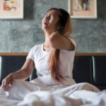 Close-up de mulher jovem sentada na cama com dor no pescoço e corpo cansado depois de acordar