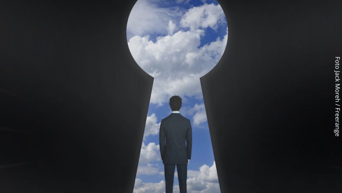 Imagem de um homem em pé de costas olhando para o céu azul por uma enorme fechadura, simbolizando um um mundo de possibilidades que uma mentalidade empreendedora oferece.