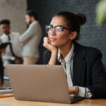 Close-up de mulher negra no escritório pensativa, sentada diante do computador, com colegas conversando ao fundo, mostrando que relacionamento interpessoal no trabalho não precisa virar amizade.