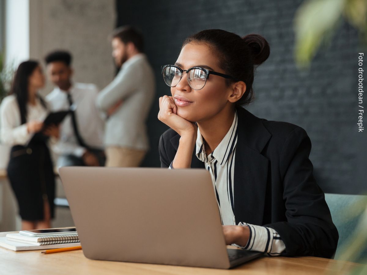 Close-up de mulher negra no escritório pensativa, sentada diante do computador, com colegas conversando ao fundo, mostrando que relacionamento interpessoal no trabalho não precisa virar amizade.
