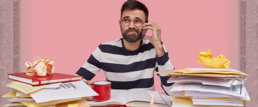 Imagem de um homem jovem em sua mesa de trabalho sobrecarregado de tarefas falando ao telefone