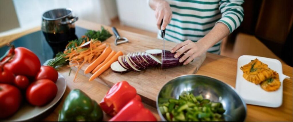 Close-up de mãos cortando um vegetal em rodelas sobre a bancada da cozinha, com outros vegetais ao redor/