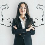 Imagem de uma mulher com os braços cruzados, à frente de dois braços musculosos abertos desenhados no fundo, simbolizando a ideia de como ser mais confiante sem precisar exibir confiança