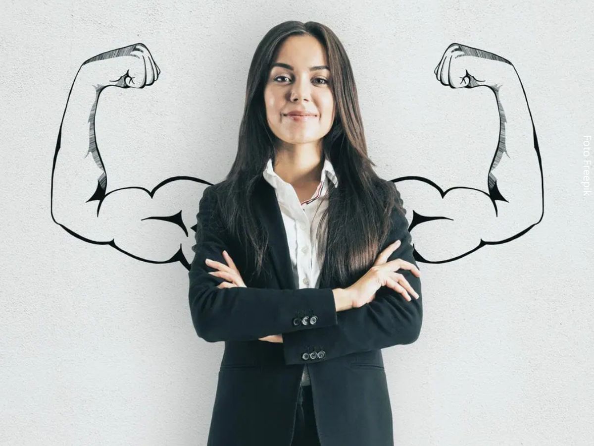 Imagem de uma mulher com os braços cruzados, à frente de dois braços musculosos abertos desenhados no fundo, simbolizando a ideia de como ser mais confiante sem precisar exibir confiança