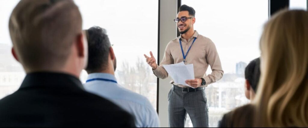 Imagem de um homem jovem de pé falando para os colegas sentados à sua frente numa reunião de trabalho