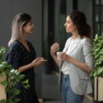 Imagem de duas mulheres conversando de pé no local de trabalho, simbolizando uma comunicação assertiva.