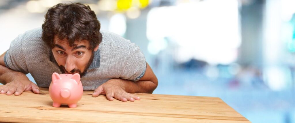 Imagem de uma homem jovem branco olhando de perto um cofre de porquinho sobre a mesa.