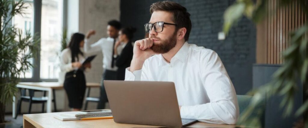 Imagem de um homem jovem branco sentado à mesa do escritório diante do computador com o olhar pensando.