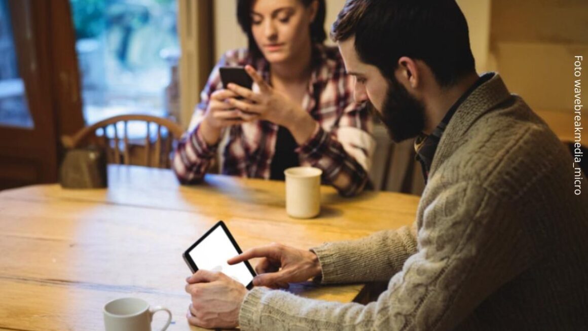 Imagem de um casal sentados à mesa de casa em silêncio com celulares na mão mantendo conversas online