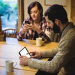 Imagem de um casal sentados à mesa de casa em silêncio com celulares na mão mantendo conversas online