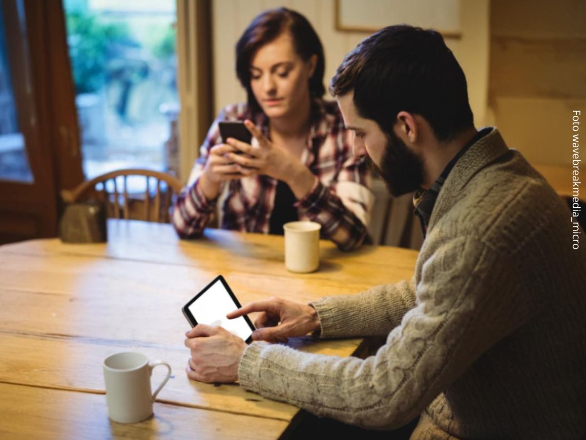 Imagem de um casal sentados à mesa de casa em silêncio com celulares na mão mantendo conversas online