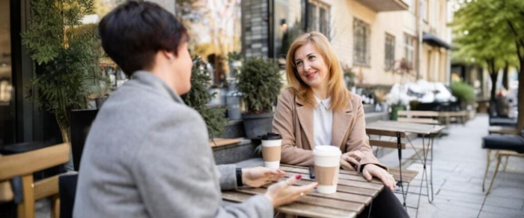Imagem de duas amigas de meia-idade sentadas à mesa de uma cafeteria conversando, tomando café e passando um tempo juntas ao ar livre