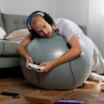 Imagem de um homem jovem jogando videogame deitado sobre uma bola de ar, sugerindo inatividade física.
