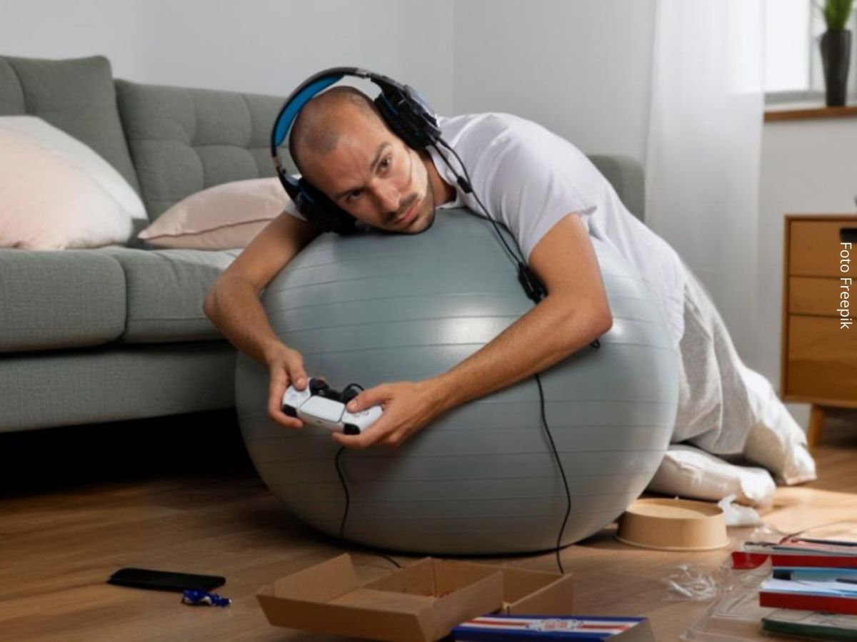 Imagem de um homem jovem jogando videogame deitado sobre uma bola de ar, sugerindo inatividade física.