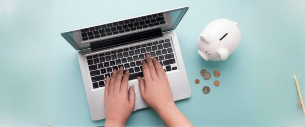 Imagem de cima de uma pessoa digitando algo no laptop, tendo ao lado um cofre em forma de porco com moedas em volta