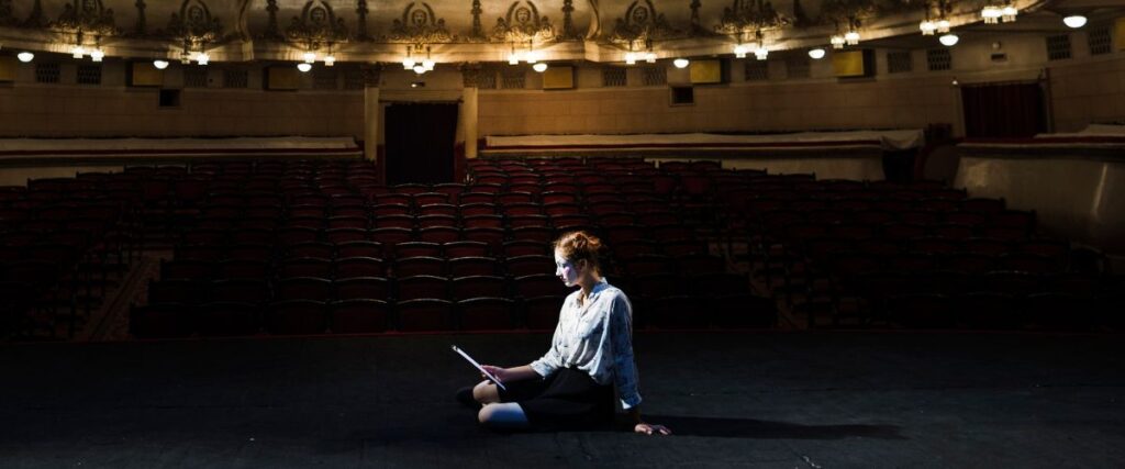 Imagem de uma atriz sentada no palco lendo o roteiro concentrada, tendo ao fundo as cadeiras do teatro vazias