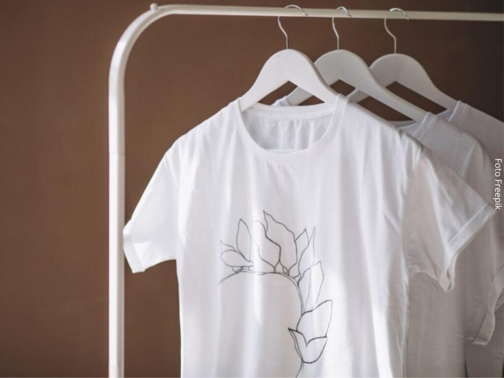 Imagem de camisetas brancas no cabide penduradas em uma arara de metal, como se fossem um mostruário de uma loja de roupa online.