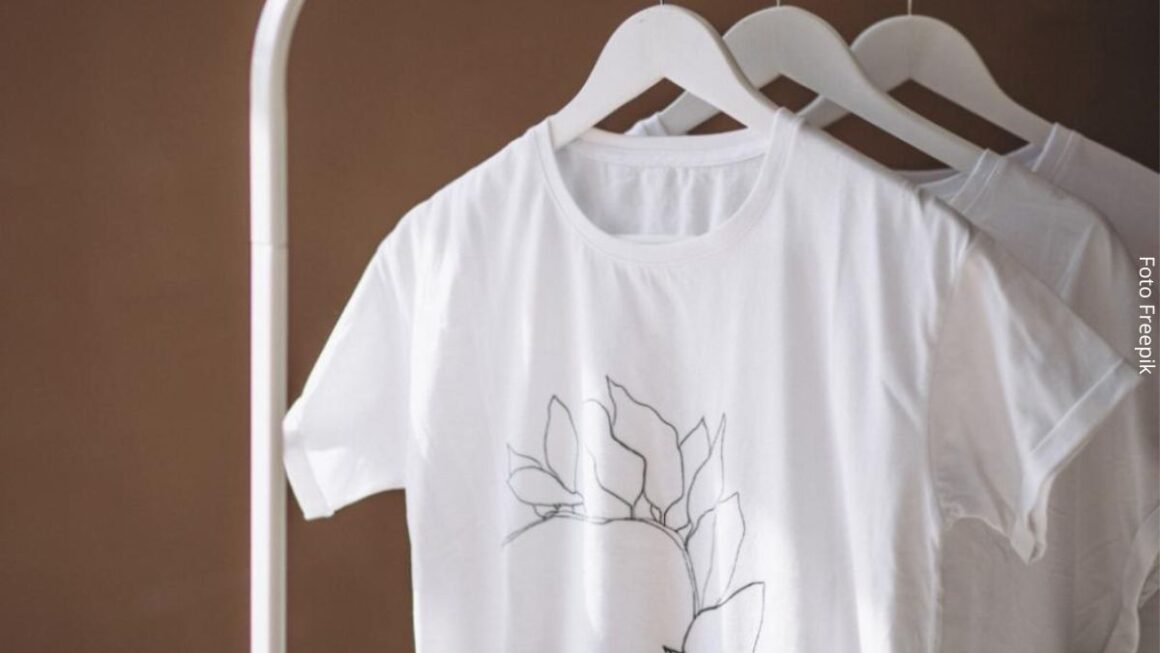 Imagem de camisetas brancas no cabide penduradas em uma arara de metal, como se fossem um mostruário de uma loja de roupa online.