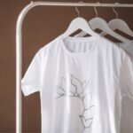 Imagem de camisetas brancas no cabide penduradas em uma arara de metal, como se fossem um mostruário de uma loja de roupa online.