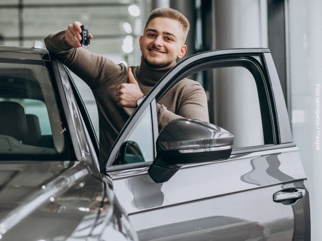 Imagem de um homem jovem na concessionária, mostrando a chave e o carro financiado que escolheu comprar