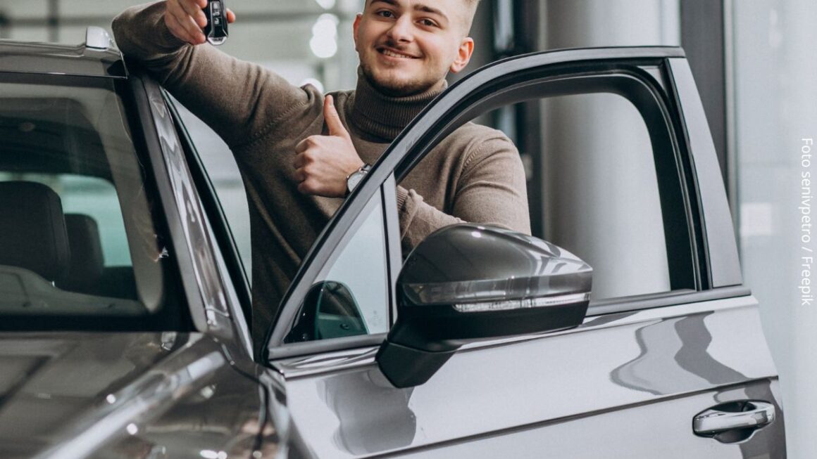 Imagem de um homem jovem na concessionária, mostrando a chave e o carro financiado que escolheu comprar