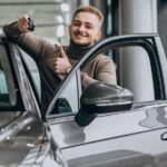 Imagem de um homem jovem na concessionária, mostrando a chave e o carro financiado que escolheu comprar