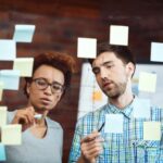 Imagem de dois líderes - uma jovem negra e um rapaz branco - no escritório, elaborando estratégias para se antecipar a problemas, exemplificando a ideia do que é ser proativo