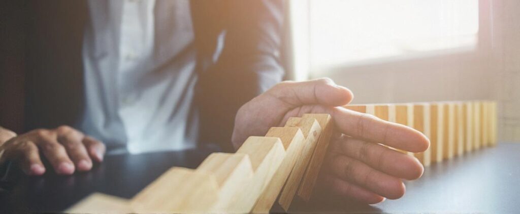 Imagem de um dominó prestes a cair, com uma peça sendo segurada por uma mão — representa a interrupção de um ciclo automático, metáfora visual do comportamento proativo.