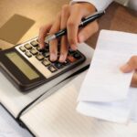 Close-up de pessoa segurando notas e fazendo contas numa calculadora, tentando se organizar para sair de dívidas.