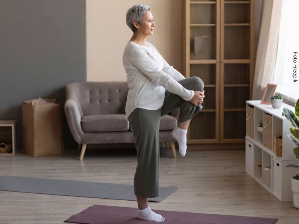 Imagem de uma mulher madura em pé sobre uma perna fazendo exercício de equilíbrio em casa.