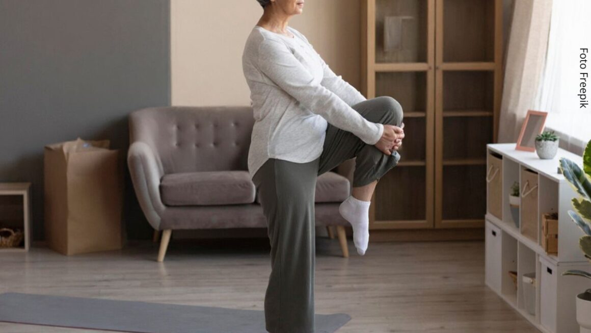 Imagem de uma mulher madura em pé sobre uma perna fazendo exercício de equilíbrio em casa.